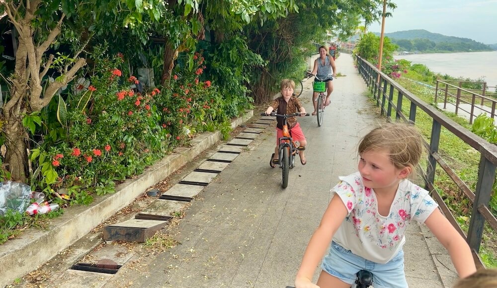 Kat, Isobel, and Henry riding bicycles along a paved boardwalk next to the Mekong River in Chiang Khan, Thailand