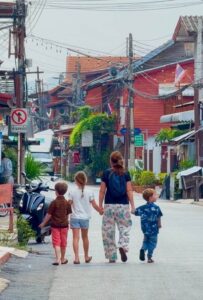Mother and three young children holding hands while walking down a peaceful street lined with traditional wooden houses in Thailand.