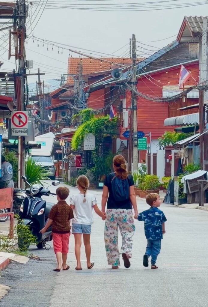 Mother and three young children holding hands while walking down a peaceful street lined with traditional wooden houses in Thailand.
