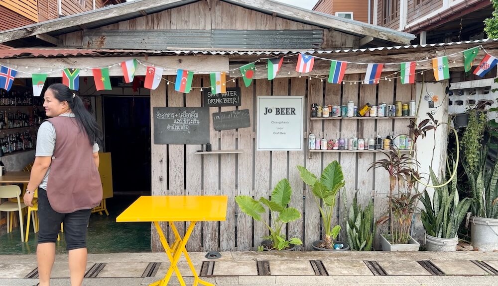Wooden rustic storefront of Jo BEER local craft beer shop adorned with international flags, shelves of craft beer cans, and potted tropical plants in Chiang Khan, Thailand.
