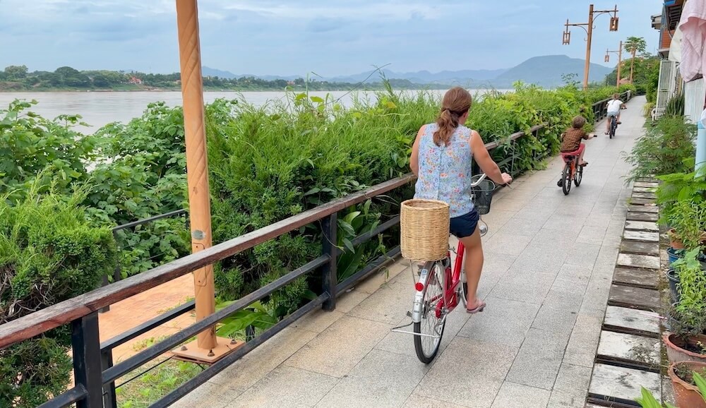 Kat riding a red city bicycle with a woven basket on the back along the Mekong River promenade in Chiang Khan with her children biking ahead.