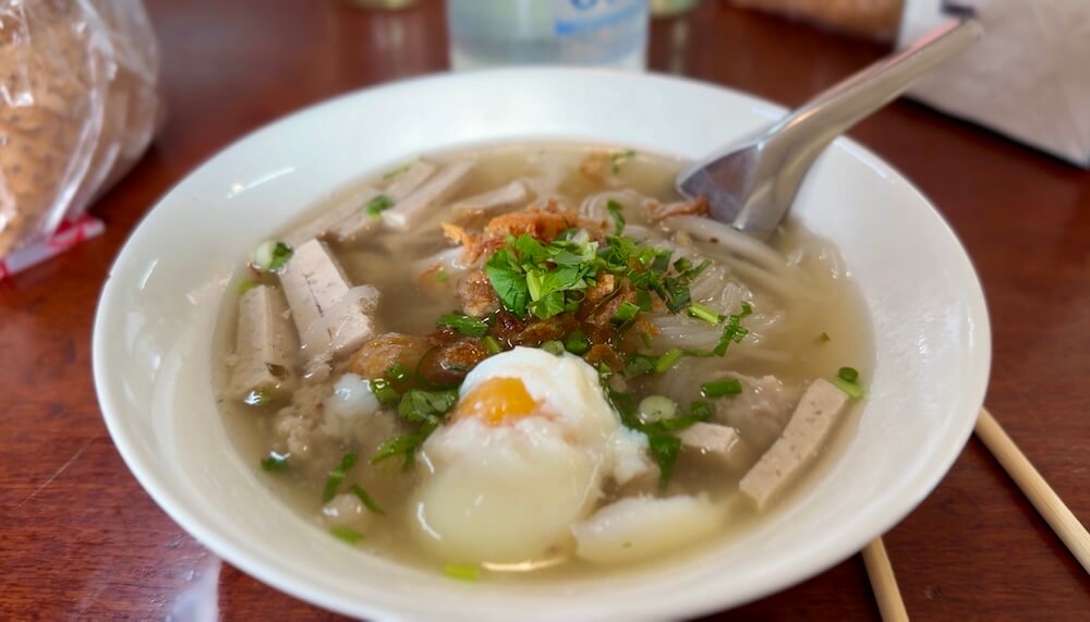 A white bowl of hot Khao Piak Sen noodle soup with chewy rice noodles, sliced pork sausage, minced pork, crispy shallots, herbs, and a soft-boiled egg in savory broth.