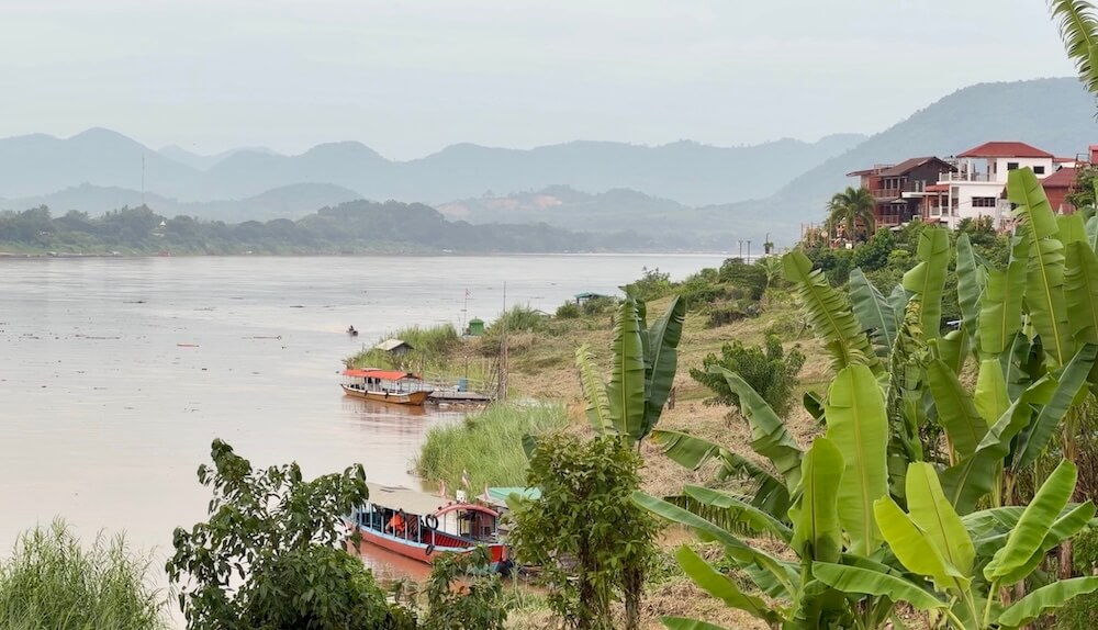 Wide view of the muddy Mekong River with wooden longtail boats docked along grassy banks framed by lush green banana trees and distant mountain hills in Chiang Khan, Thailand.