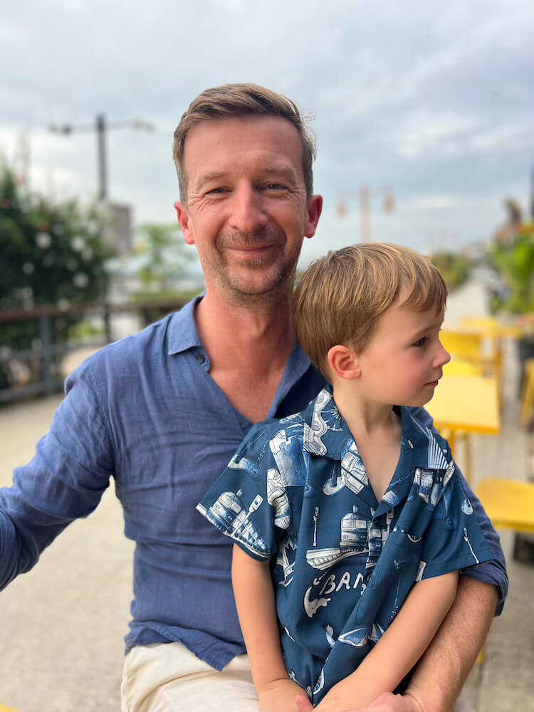 A father smiling at the camera while holding his young son on his lap at an outdoor café seating area in Thailand.